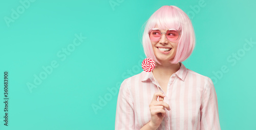 Banner of young woman in shirt  pink wig and glasses  holding lollipop in hand  biting lip and looking away  isolated on mint blue background