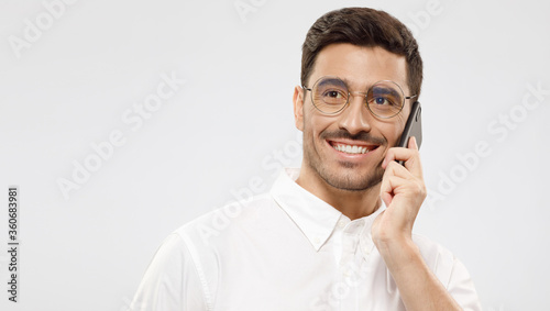 Horizontal banner of young business guy in shirt and glasses, answering phone call with happy smile, looking aside, isolated on gray background with copy space