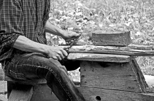 This man is using a pioneer draw knife on wood, used to remove bark from timber in this vintage, black and white lifestyle image.
