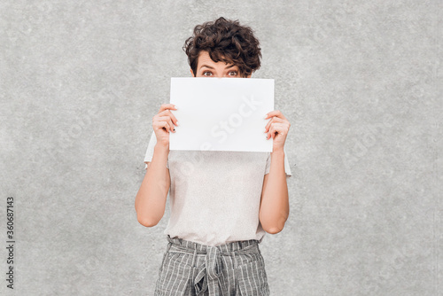 Young pretty woman with creative hairstyle smiles and hide behinde blank piece of paper in her hand. photo