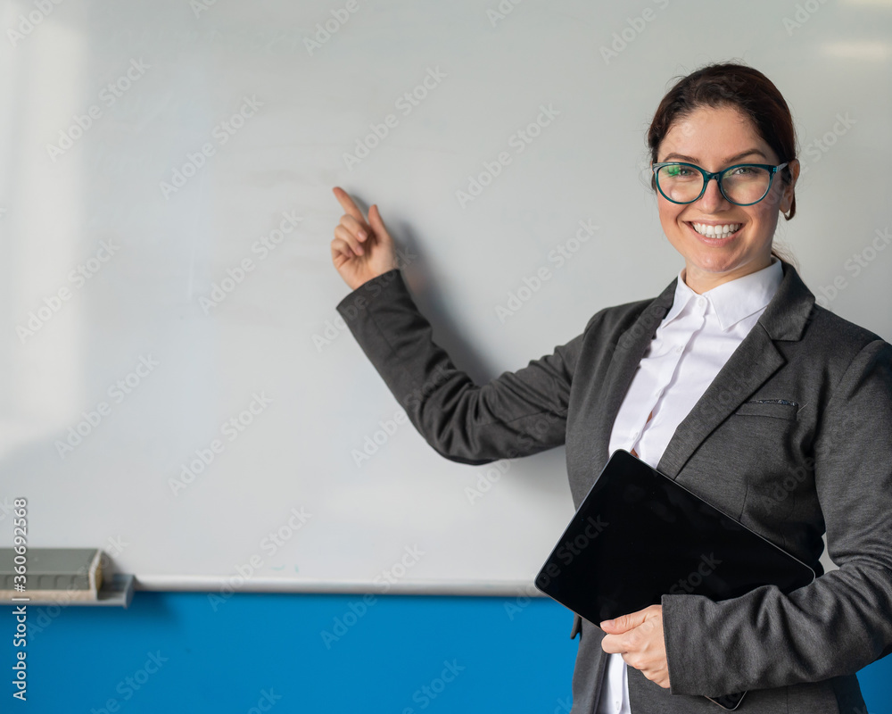 Beautiful red-haired woman in a suit with a digital tablet in her hands. A smiling female teacher with glasses stands in a school class and points a finger at a white board. Educational institution. Beautiful red-haired woman in a suit with a digital tablet in her hands. A smiling female teacher with glasses stands in a school class and points a finger at a white board. Educational institution.