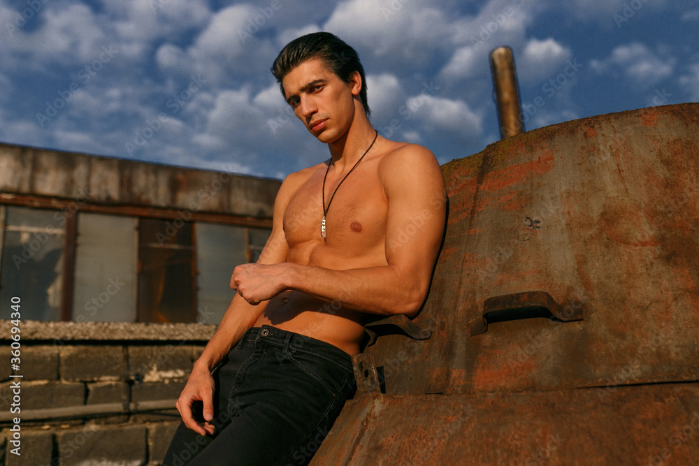 Portrait of a confident athletic young man with naked torso posing on the roof at the sunset background.