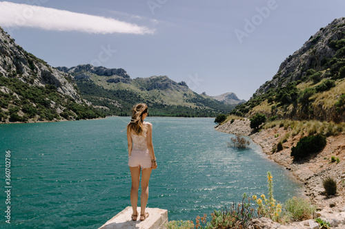 Young girl stands on pier near mountain lake with her back to the frame. Her pink summer clothes and hair develop on a winter. Romantic panorama  © Stella