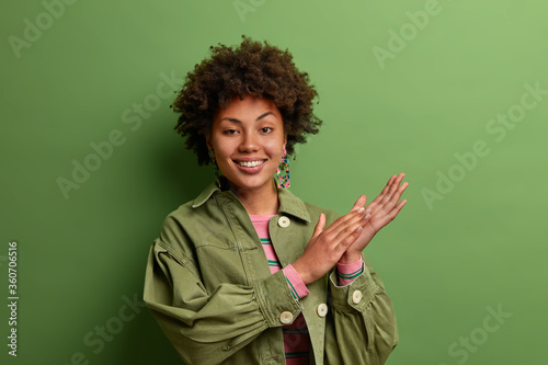 Positive mysterious woman smiles toothily, rubs palms with intention to make dream real, enjoys good result of work, wears fashionable green jacket, poses indoor, has optimistic glad expression