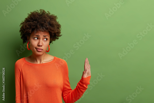 Serious displeased dark skinned woman shows palms sideways in prohibition gesture, refuses something, says its not for me, dressed in bright clothes, isolated on green background, rejects offer photo