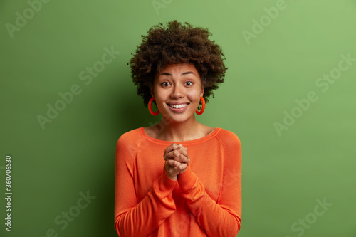 Young smiling woman with Afro hair, keeps hands pressed together, looks with friendly pleased look at camera, anticipates something good happen, awaits positive answer from you, models indoor