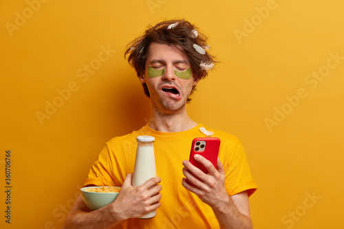 Tired male student studied all night before exam, has lack of sleep, yawns with drowsy expression, texts message on smartphone, holds bottle of milk and cornflakes, going to have breakfast in morning © wayhome.studio 