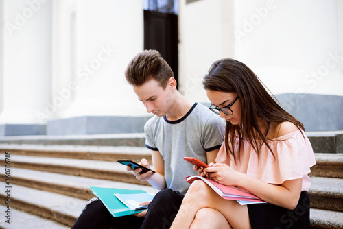 Two students sitting on stairs of university and watching something in smartphones, holding folders and copybooks, looking concentrated