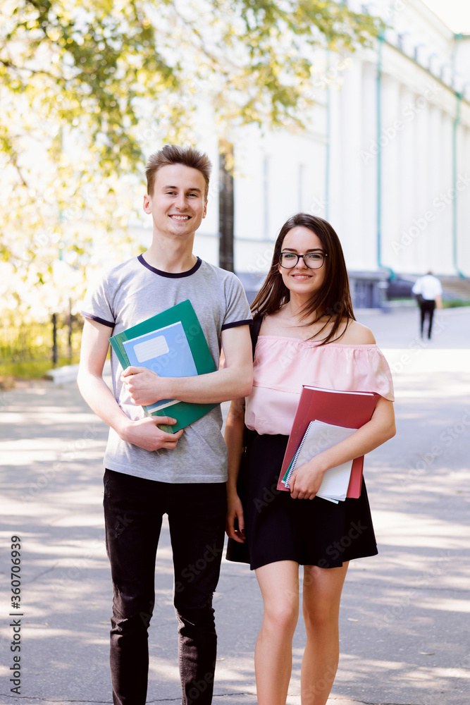 Full length portrait of two cheerful students standing in college holding learning materials dressed in casual clothes