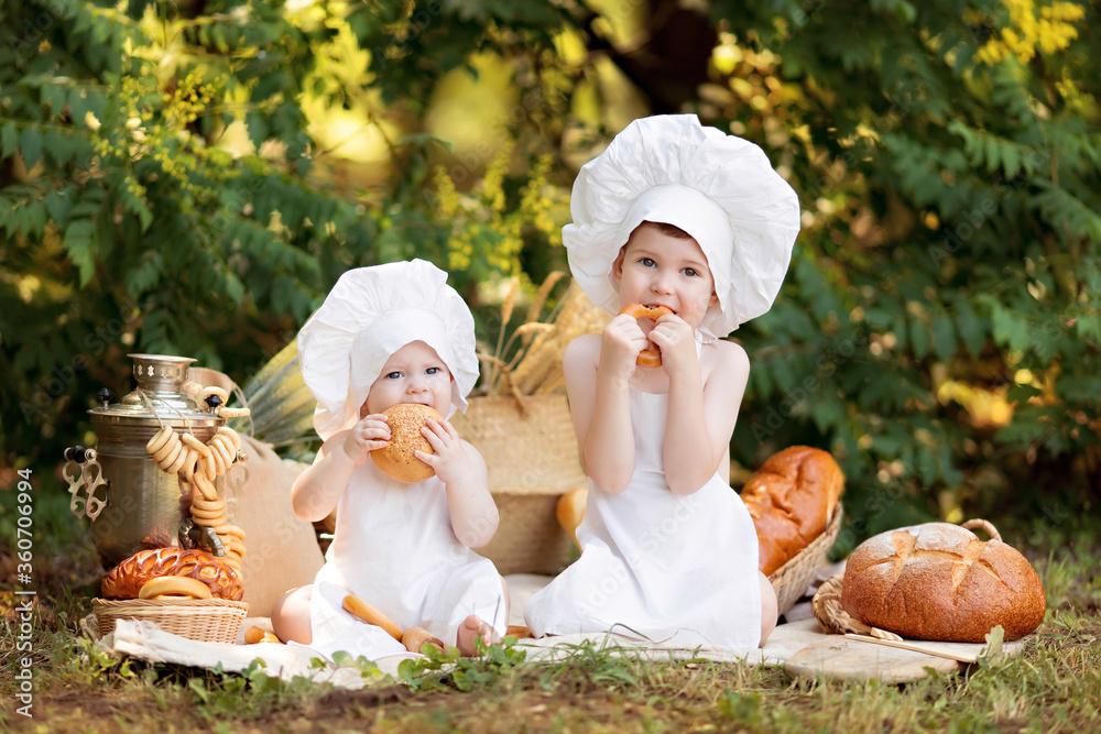 Healthy eating concept. Happy little girl and boy cook outdoors on a sunny summer day. Toddler baker on a picnic eats bread and bagels in a white apron and hat
