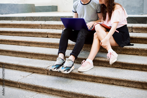 Male and female students sitting at the stairs of university, using laptop with pleasant smiles on their faces. Students and education concept.