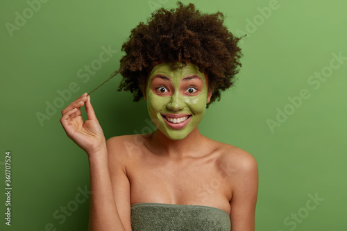 Positive Afro American woman touches curl, has frizzy hair, smiles broadly and shows snow white teeth, wrapped in bath towel, applies green moisturizing mask on face, isolated on green background