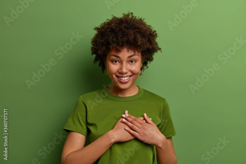 Smiling cute female with dark curly hair, makes gratitude gesture, presses hands to heart, swears or promises, has sincere look, white perfect teeth, wears bright green t shirt, poses indoor