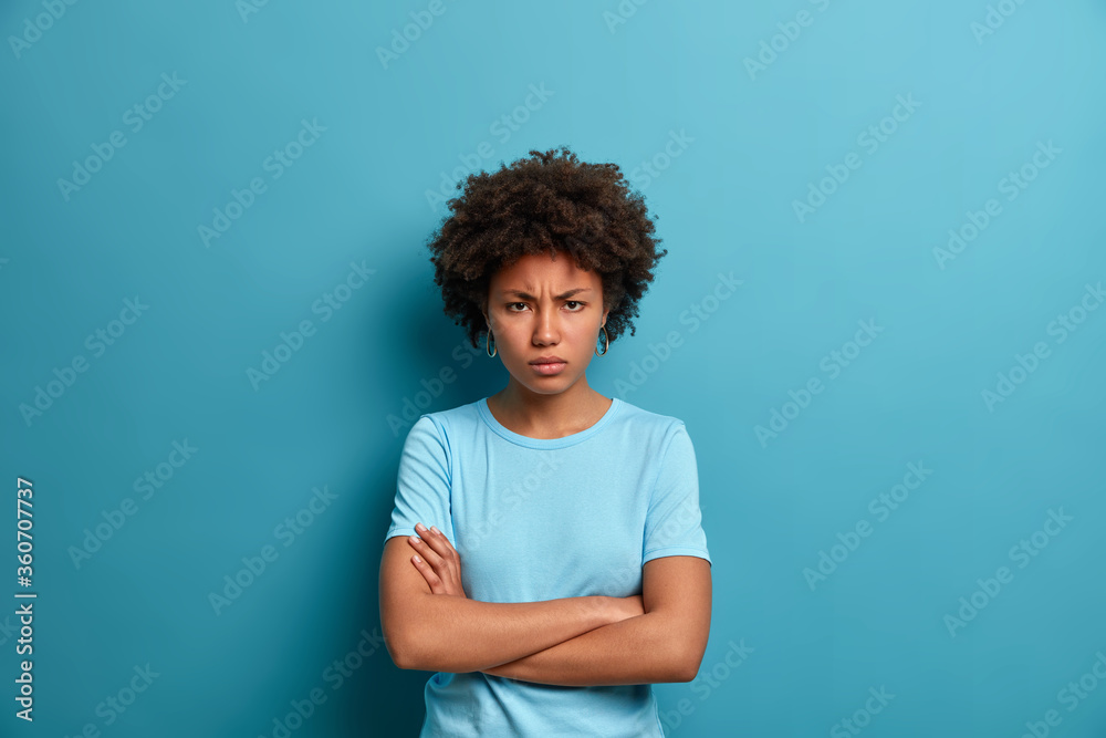 Horizontal shot of resentful offended woman crosses hands over body, looks at camera with angry gaze and sullen expression, being angry with someone, has formidable look, poses over blue studio wall Horizontal shot of resentful offended woman crosses hands over body, looks at camera with angry gaze and sullen expression, being angry with someone, has formidable look, poses over blue studio wall