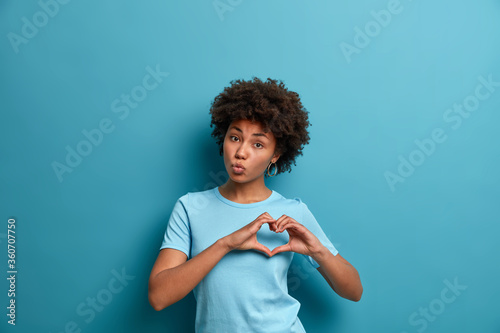 Romantic beautiful curly haired young woman expresses feelings with amorous heart gesture, demonstrates symbol of affection and devotion, keeps lips folded, poses against blue studio background