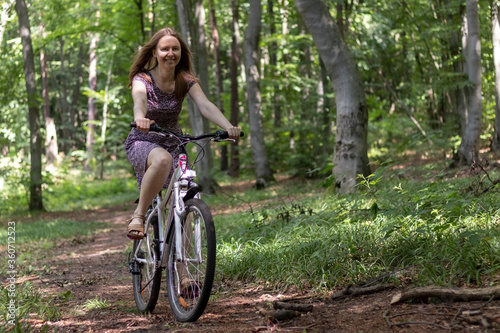 A young woman in a light dress rides a bicycle through a forest.
