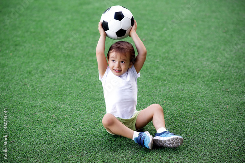 Fototapeta Naklejka Na Ścianę i Meble -  Toddler boy in sports uniform sitting on green footall field holding soccer ball above head. Little child playing with ball on grass. Active childhood and outdoor games for children concept