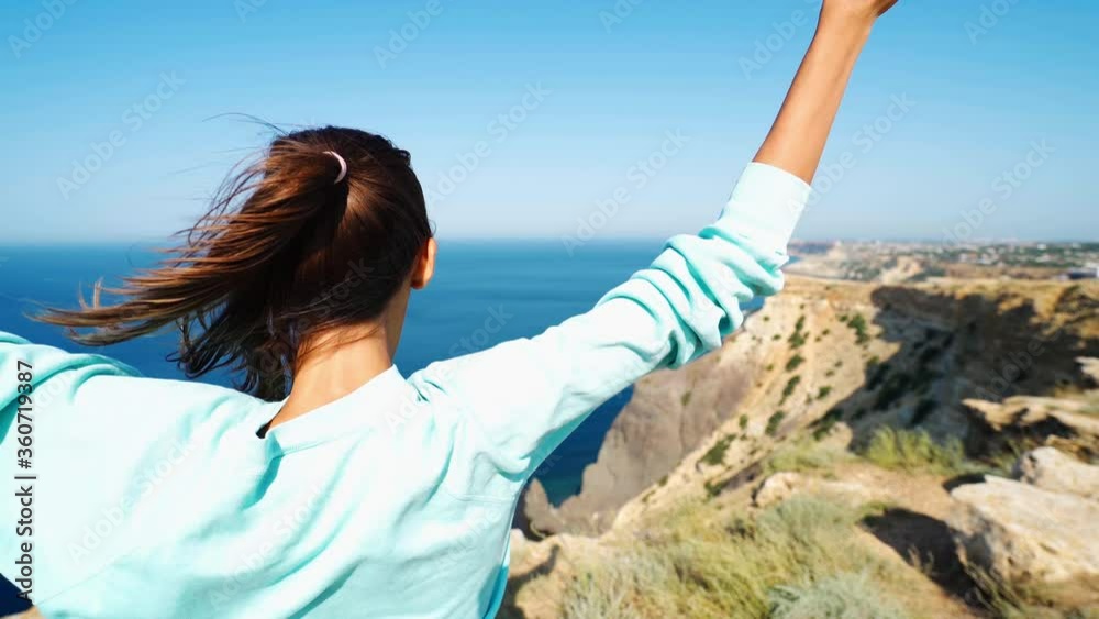 rear view happy young Woman traveller with arms raised, celebrating scenic seascape enjoying vacation travel on top of mountain looking at sea view. Hiker girl lifting arm up hair wind blowing. rear view happy young Woman traveller with arms raised, celebrating scenic seascape enjoying vacation travel on top of mountain looking at sea view. Hiker girl lifting arm up hair wind blowing.