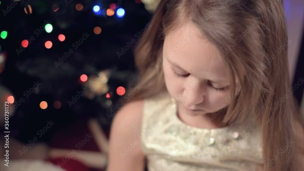 A little girl in a festive dress against the backdrop of festive lights and a Christmas tree sadly looks somewhere down the blur and movement to focus. The concept of a spoiled holiday A little girl in a festive dress against the backdrop of festive lights and a Christmas tree sadly looks somewhere down the blur and movement to focus. The concept of a spoiled holiday