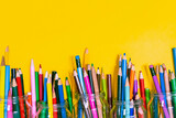 Many colored pencils are lying on the bottom of the bright yellow background. The place for the title is an empty sheet. Top view.