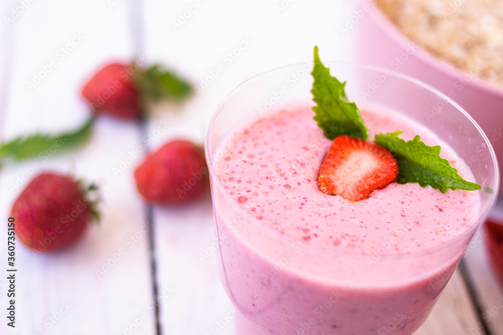 Cocktail with strawberries and milk in a glass decorated with mint and strawberries. Nearby is a pink cup with Hercules flakes, and loose strawberries on a white wooden background. Cocktail with strawberries and milk in a glass decorated with mint and strawberries. Nearby is a pink cup with Hercules flakes, and loose strawberries on a white wooden background.