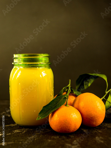 Fresh tangerine juice in a glass jar on a black background. Whole tangerines with green leaves lie nearby. Healthy eating concept