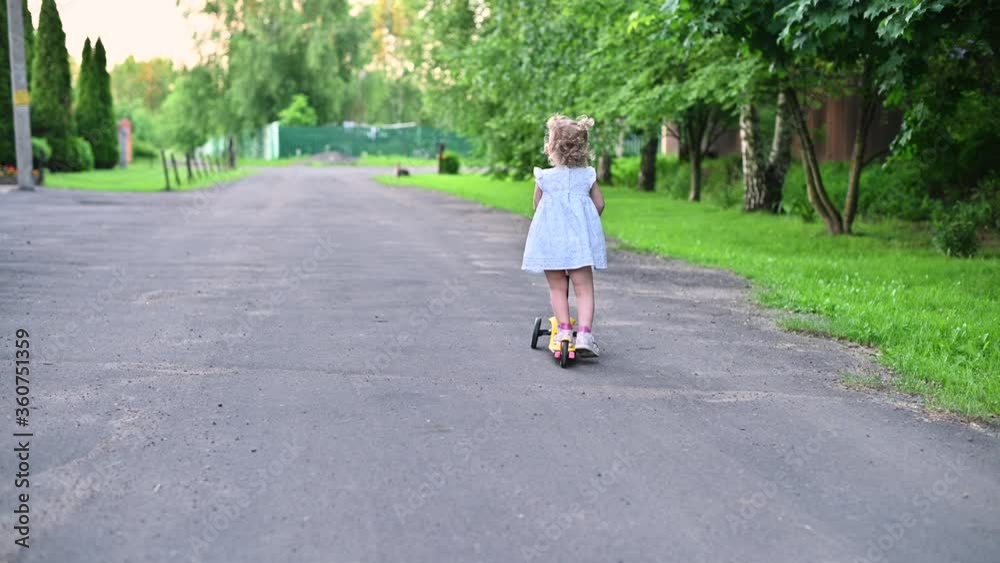 Little girl leaves the camera on a children's scooter and looks back. Walking on a summer evening in the village Little girl leaves the camera on a children's scooter and looks back. Walking on a summer evening in the village
