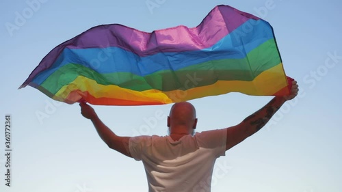 Homosexual bold man with rainbow gay flag while parade on background of blue sky. LGBTQI, Pride Event, LBGT Pride Month, Gay Pride Symbol