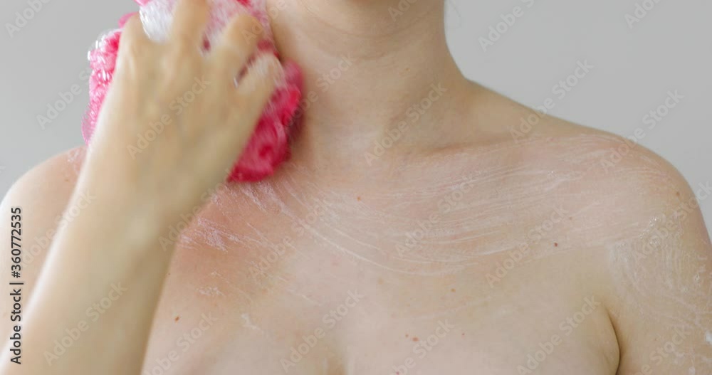 Woman is washing rubbing shoulders and chest with foam using pink loofah, front view. Woman is taking a shower applying organic gel on skin on grey background. Massaging her body with bath sponge. Woman is washing rubbing shoulders and chest with foam using pink loofah, front view. Woman is taking a shower applying organic gel on skin on grey background. Massaging her body with bath sponge.