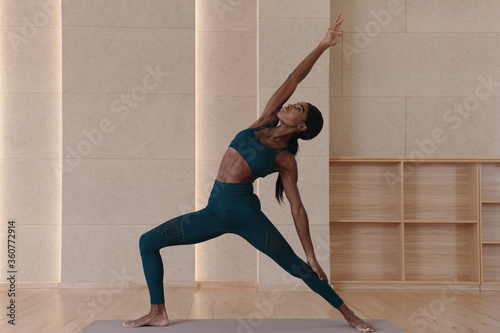 Young black woman practicing yoga, doing Exalted Crescent Lunge Twist on the Knee exercise, indoor full length, studio background