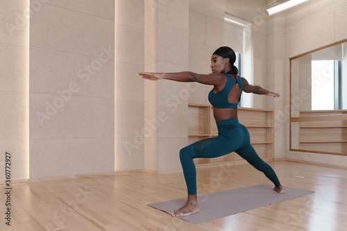 Young black woman practicing yoga, doing Virabhadrasana 2 exercise, Warrior two pose, indoor full length, studio background