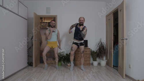 Brutal men in women's shorts train with dumbbells at home in conditions of self-isolation, mimicking women. They make faces and have fun in the frame and perform exercises asynchronously. The frame photo