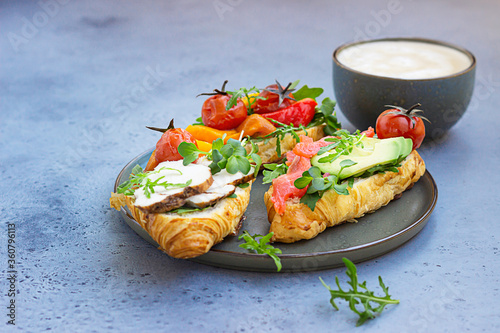 Breakfast or lunch with coffee and different croissant sandwiches with grilled pepper, tomatoes, smoked salmon, turkey, avocado and arugula served with micro green. Grey stone background.