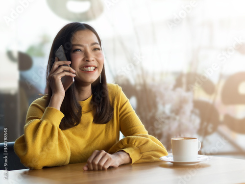 Woman talking on mobile phone and smiling happily while having hot coffee in the coffee shop.