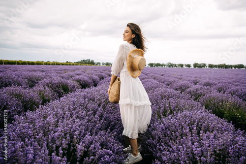 Young romantic woman standing in lavender field, wearing a bohemian white maxi dress and a straw hat.