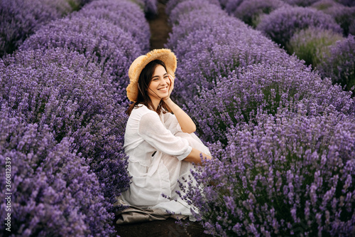 Young brunette woman sitting between rows of lavender bushes, smiling, wearing a bohemian white dress and a straw hat.