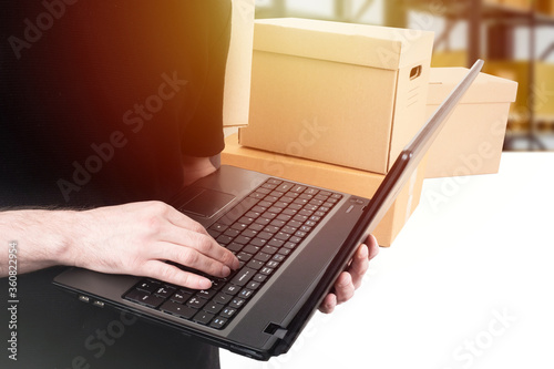 Goods accounting. Accounting of the goods. Men's hands with a laptop on the background of cardboard boxes. The man keeps track of the product. A person is engaged in sending parcels.