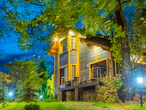 Vacation home. Concept - rest in a country cottage. House in the nature. Two-story house at night. Concept - sale of real estate. There are a lot of trees around cottage. Lanterns illuminate the area