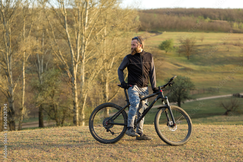 Sports brutal bearded guy on a modern mountain bike. Cyclist on the green hills in the spring.