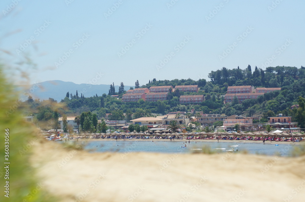 A crowded beach on a hot summer day in Greece. People are relaxing under umbrellas. Behind them are hotels on a green hill. On the background are mountains. 
