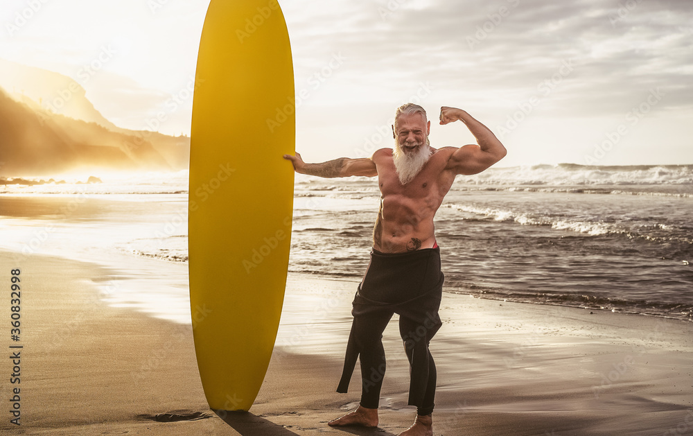 Happy fit senior having fun surfing at sunset time - Sporty bearded man training with surfboard on the beach - Elderly healthy people lifestyle and extreme sport concept - obrazy, fototapety, plakaty 