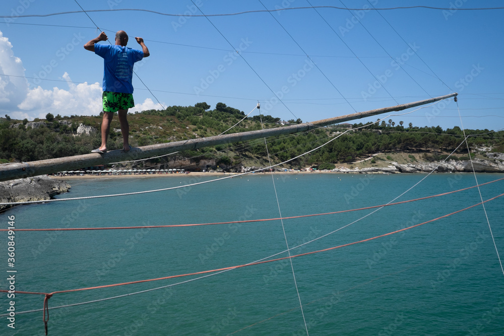 Naklejka premium Unrecognizable fisherman walks over a wooden pole and holds onto the ropes of a traditional fishing trabucco at the beach of Vieste, Italy