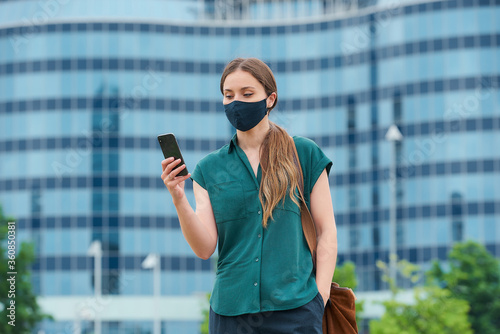 A woman in a navy blue medical face mask to avoid the spread of coronavirus in the center of the city. A girl with reading the news on smartphone thrusting a hand into a pocket of trousers in downtown