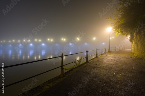 Fototapeta Naklejka Na Ścianę i Meble -  Moody night view of empty street and barrier along the Loire River, illuminated by a street lamp, with a lit bridge appearing in the fog in Saumur, France