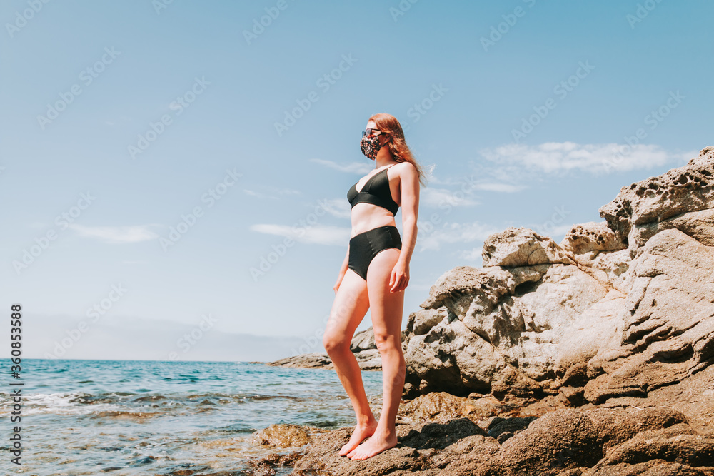 Woman in bikini and stylish face mask at empty beach, new normal rules, beach vacation. Life after pandemic, obligatory use of face mask in public spaces. Summer during covid, new normal concept
