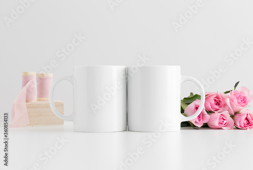 Two mugs mockup with a bouquet of pink roses and silk ribbons on a white table.