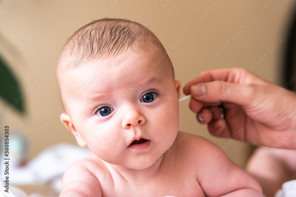Cute little caucasian baby boy looking at camera while ear cleaning by his parent. Happy baby enjoying bath and hygiene care time at home.