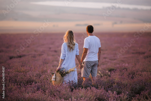 Loving couple admires the beautiful sunset in the lavender field. The guy and the girl are standing with their backs and holding hands.