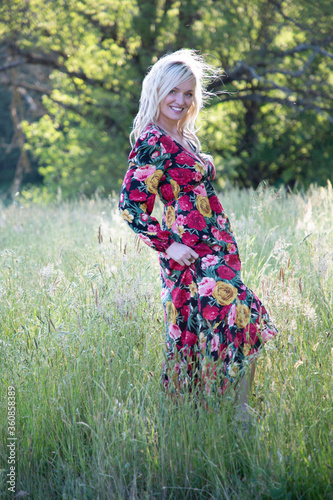 Donna bella e bionda con un vestito lungo a fiori in un campo con l'erba alta e gli alberi sullo sfondo