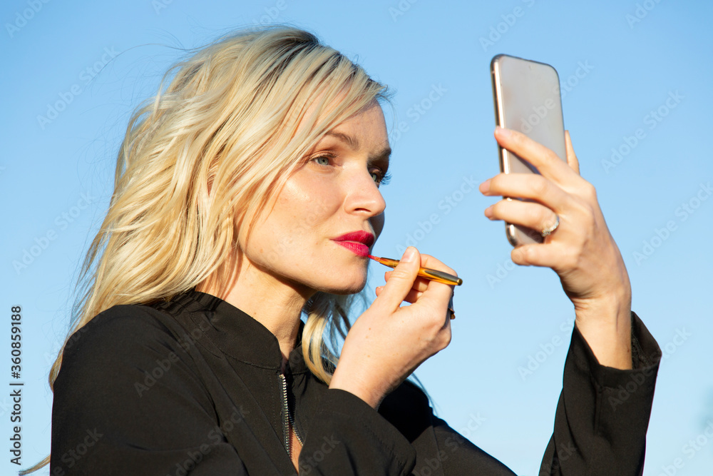 Giovane donna bella e bionda che si trucca usando il telefonino come uno specchio, vestita di nero illuminata dalla luce del sole, primo piano Giovane donna bella e bionda che si trucca usando il telefonino come uno specchio, vestita di nero illuminata dalla luce del sole, primo piano